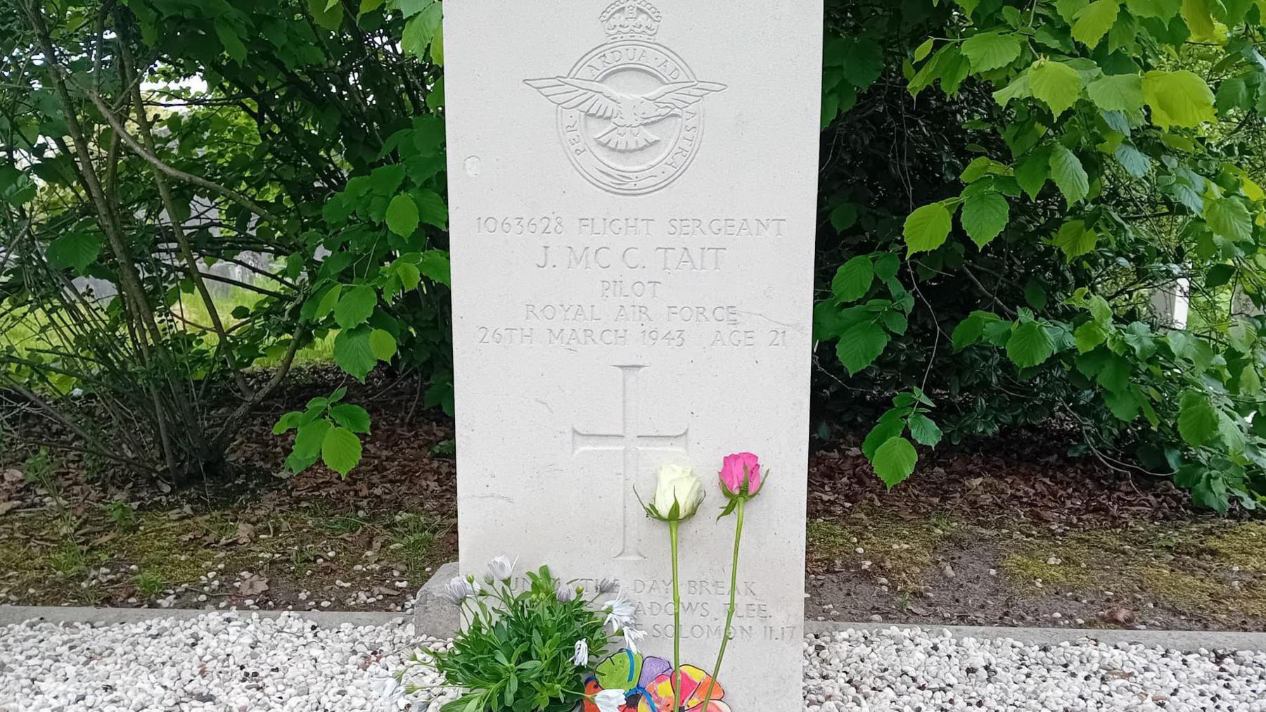 An image of a grave, the headstone reads 'Flight Sergeant J.MC C. Tait, Pilot, Royal Air Force, 26th March 1943, Age 21.' Two roses, one white and one pink and a plant have been placed at the bottom.