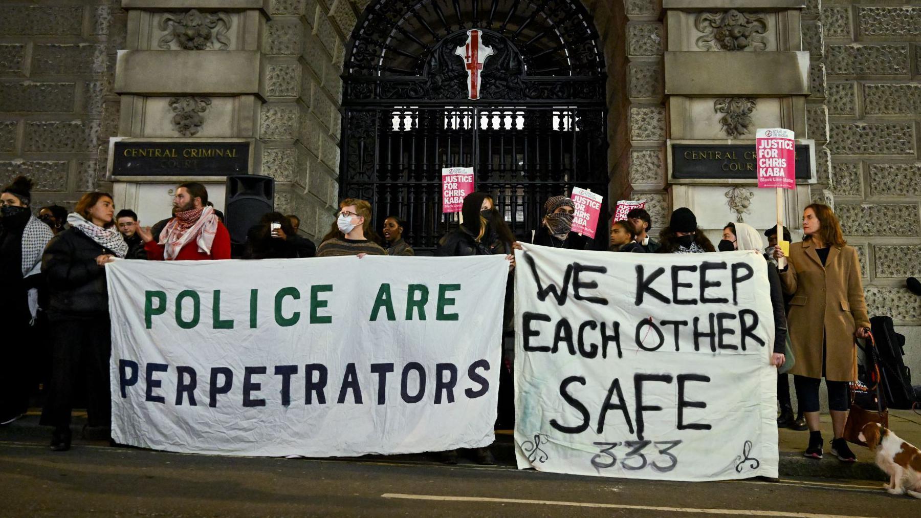 A group of about 30 protestors standing peacefully and holding home-made banners with "police and perpetrators" and "we keep each other safe" 