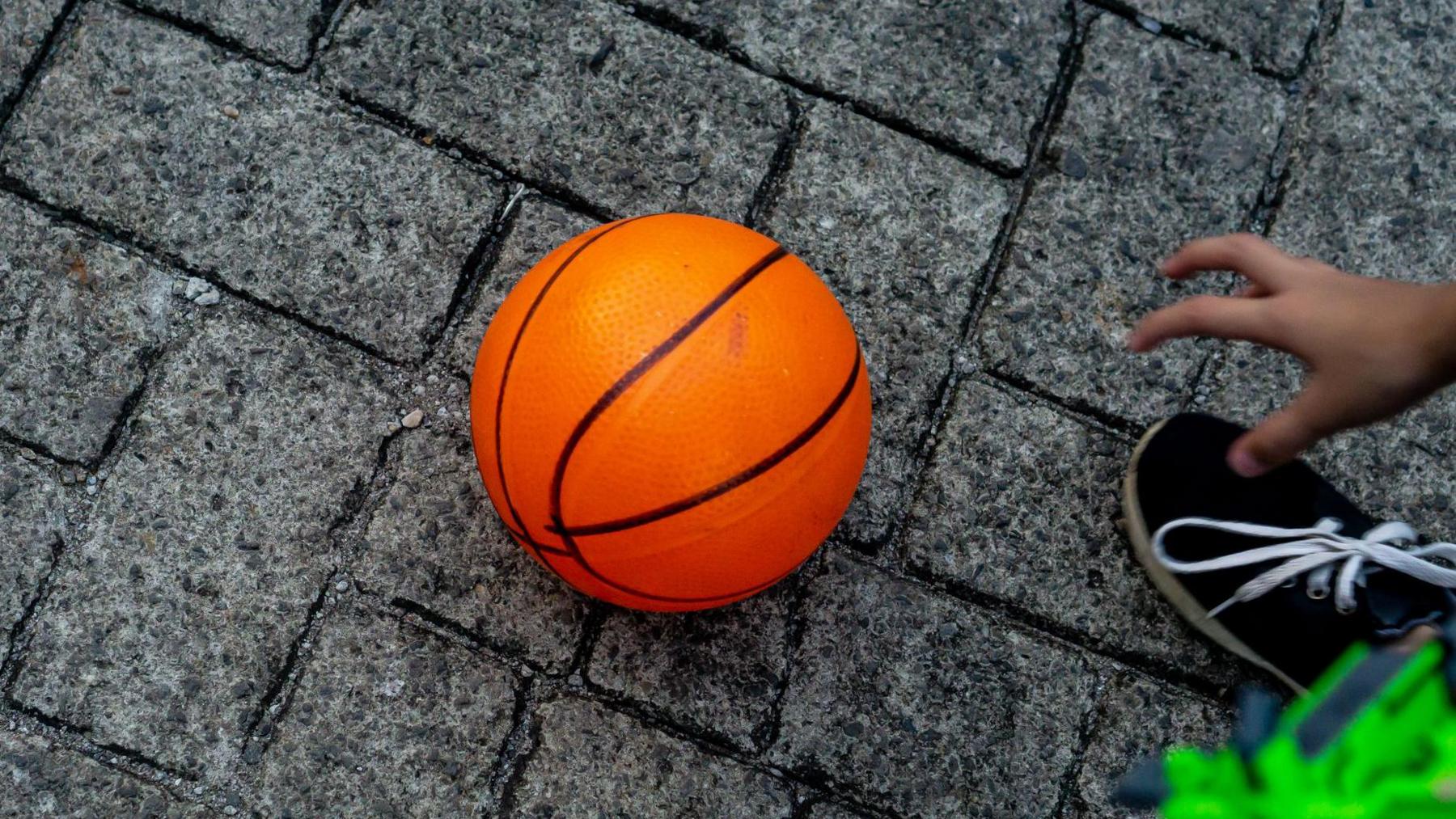 A hand is seen reaching towards an orange basketball which is lying on a grey brick pavement.