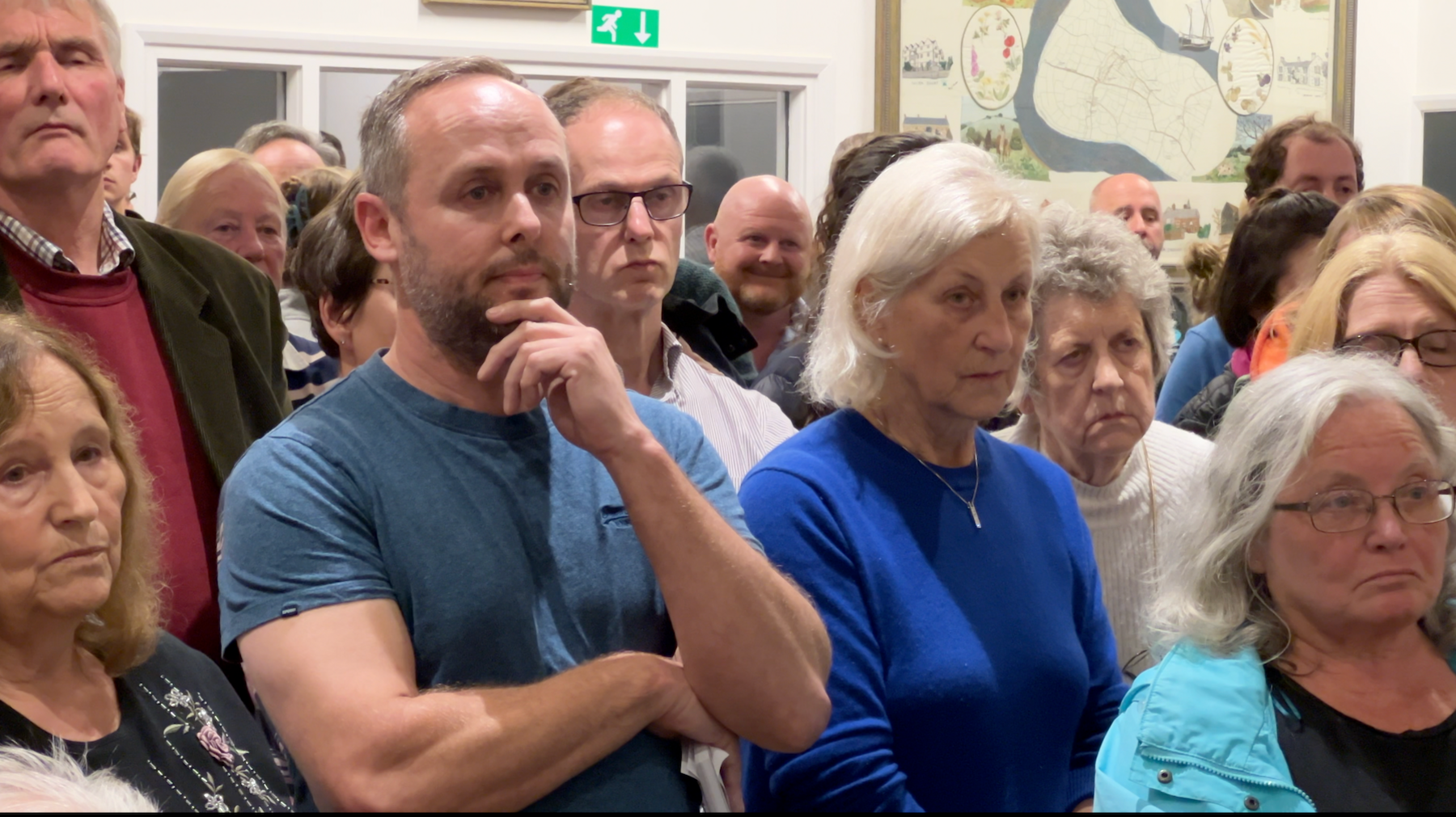 Dave Seal stands in the public meeting, his hand to his chin as he listens. He wears a blue T shirt, and is surrounded by others in the crowd.