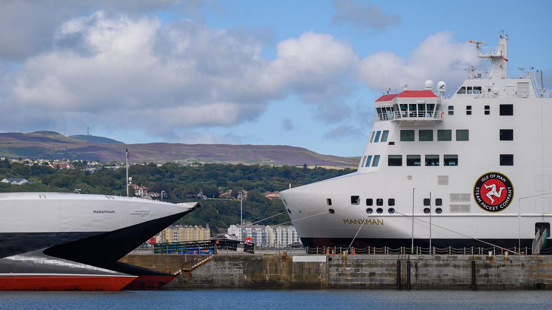 Two white, red and navy passenger ferries pointing in the same direction whilst docked on either side of a harbour, appearing as though the noses of their bows are close together. 
Hills are in the background. 