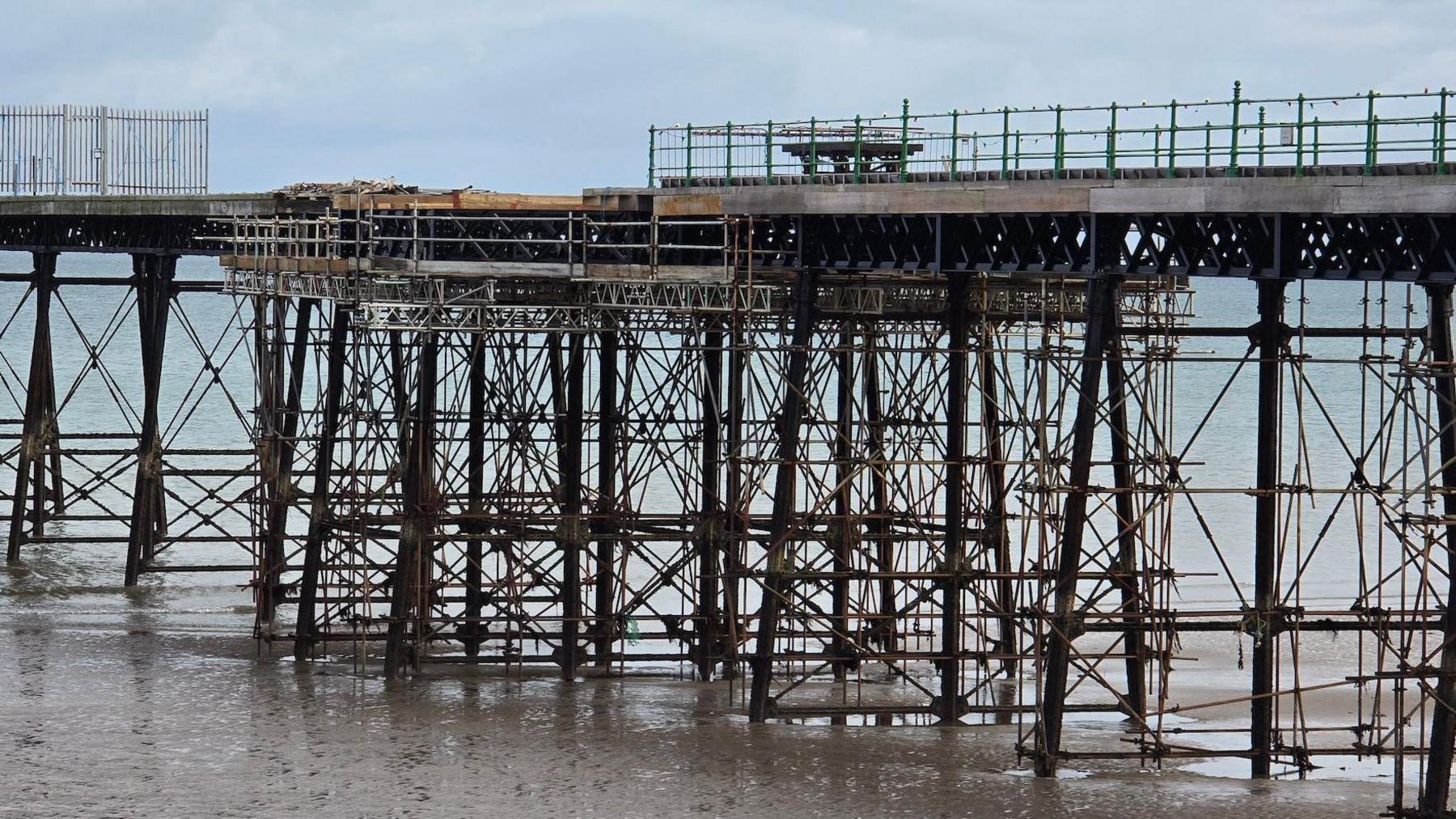 A close-up of a section of the iron pier, with the central section surrounded by scaffolding. There is fencing on the platform. At the base, the large rods meet a sandy beach and a calm sea on a cloudy day.