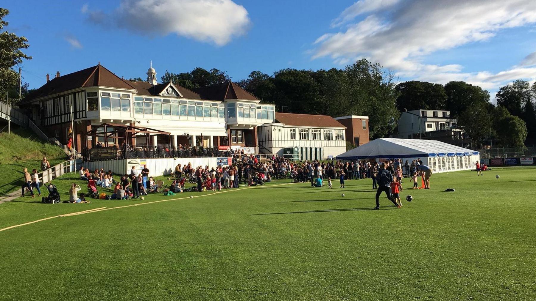 A Victorian white clubhouse with lots of people gathered outside. There is a marquee to the right.