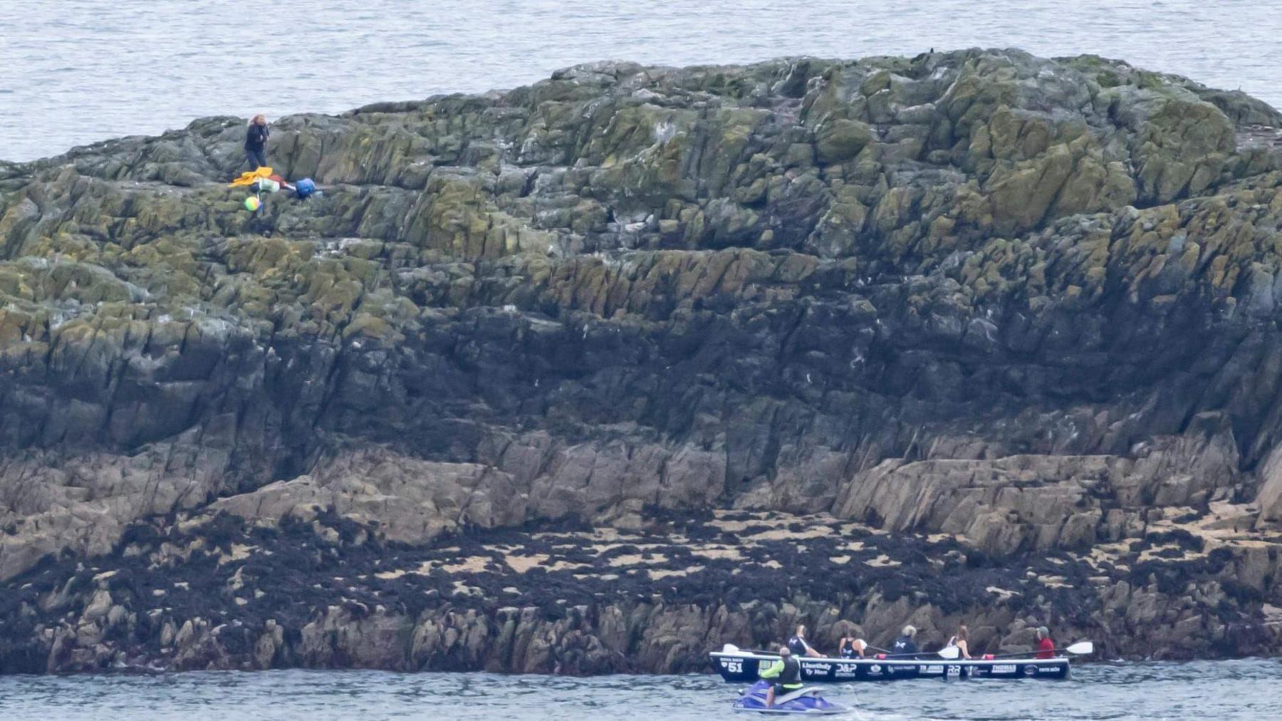 A woman, Di Mills, is seen standing on a small rocky island after being dropped off by helpers seen sitting in a small boat in the surrounding waters