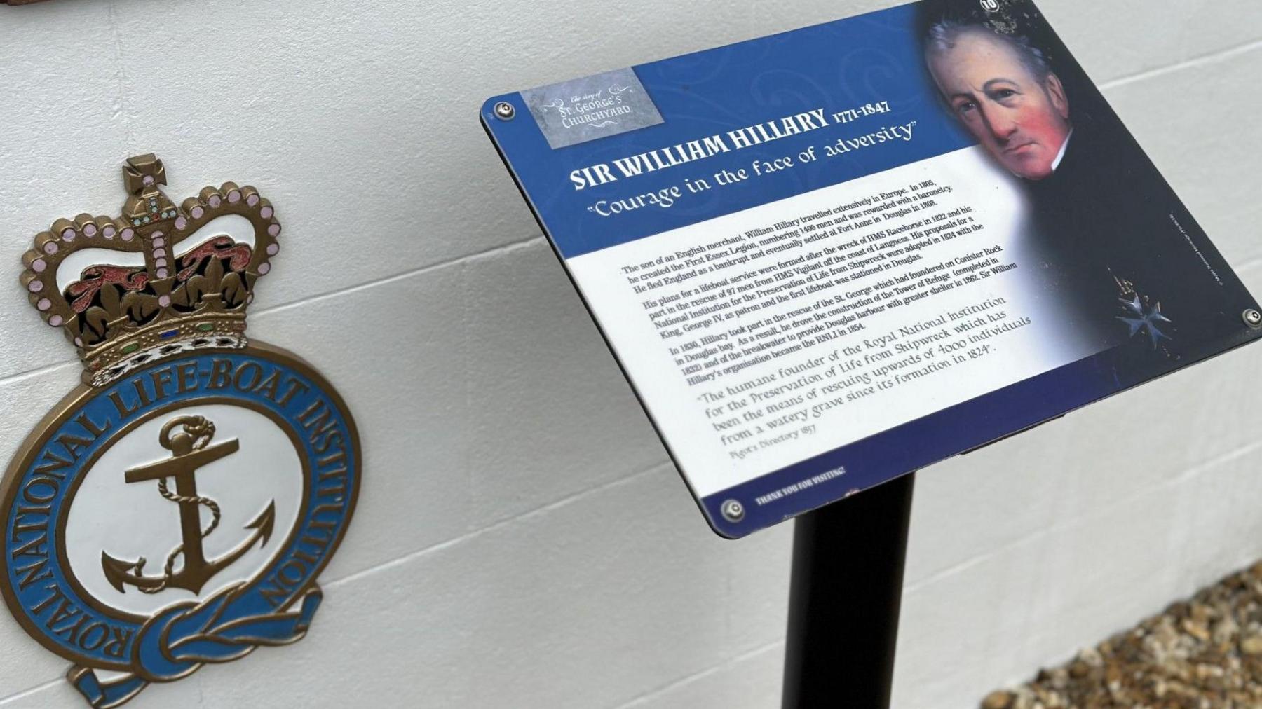 An emblem on the side of Sr William Hillary's white painted tomb featuring a crown on top if a blue circle with an anchor in the middle of it and the word Royal National Lifeboat Institution written around it. There is also a board featuring a picture of Sir William and information about him.