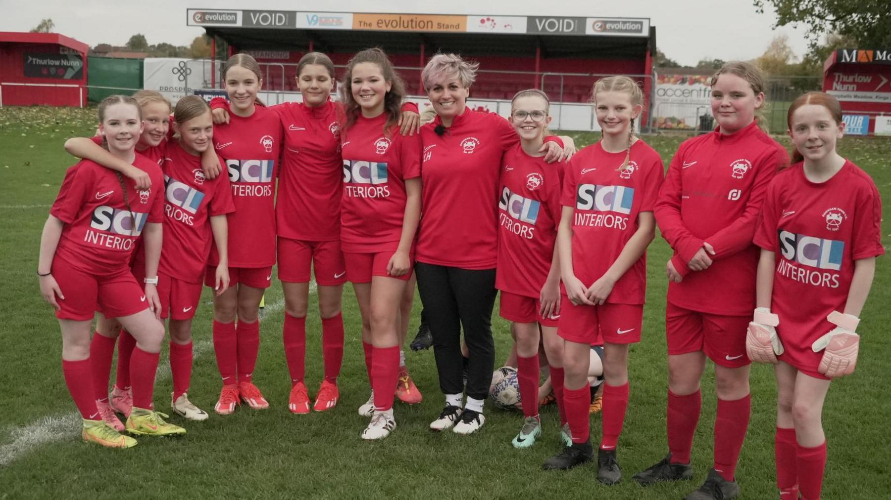 Female coach surrounded by girls wearing red tops and shorts and football boots.