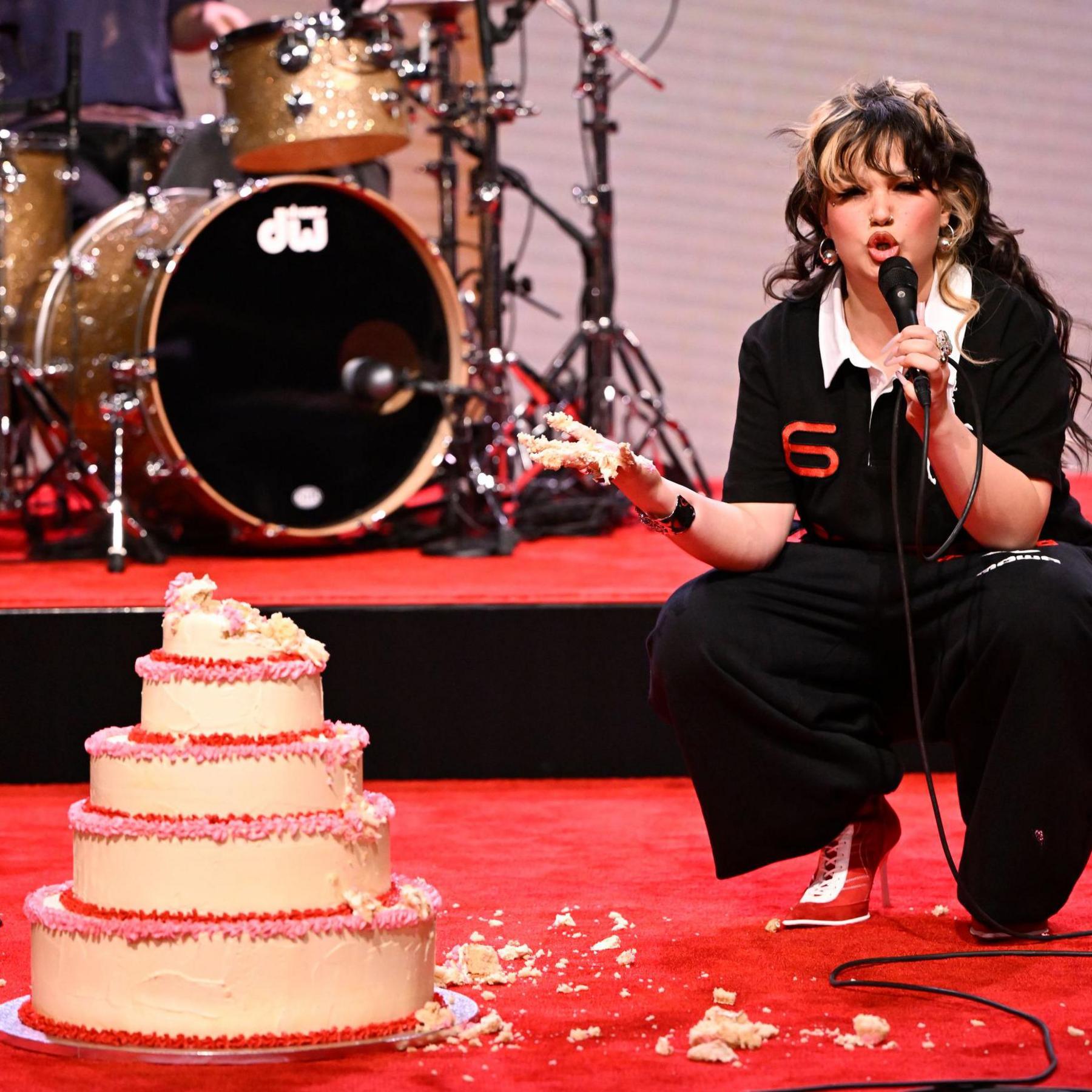 Lola Young performing on the Jimmy Fallon show. She wears black trousers and a black polo shirt and crouches next to a five-tiered cake with cream, red and pink frosting. She's gouged out the top tier and her cake is covered in cake. She's performing on a red carpet and behind her on set is a golden drum kit.