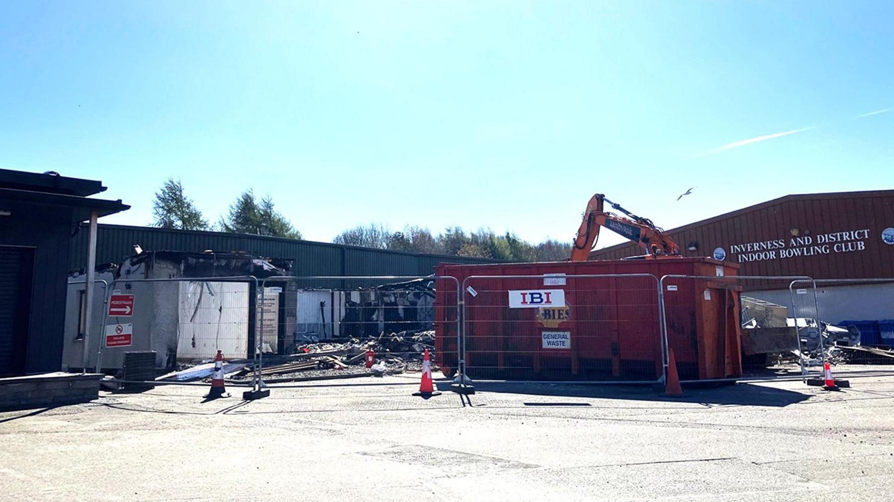 Much of the fire damaged have been removed. There is large wire fencing around the site. There is a large red skip and just visible the arm of an excavator.
