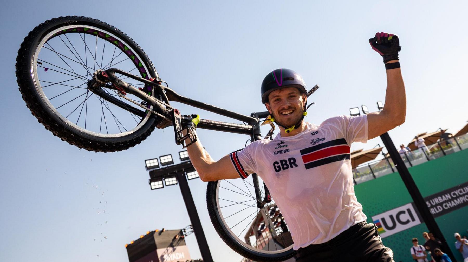 Charlie Rolls smiles at the camera as he holds one of his arms up in the air in celebration. In his other hand he holds his bike up in the air. He wears a black helmet with a white top that has GBR written on the chest and he wears black shorts as well as gloves. He has some stubble on his face.
