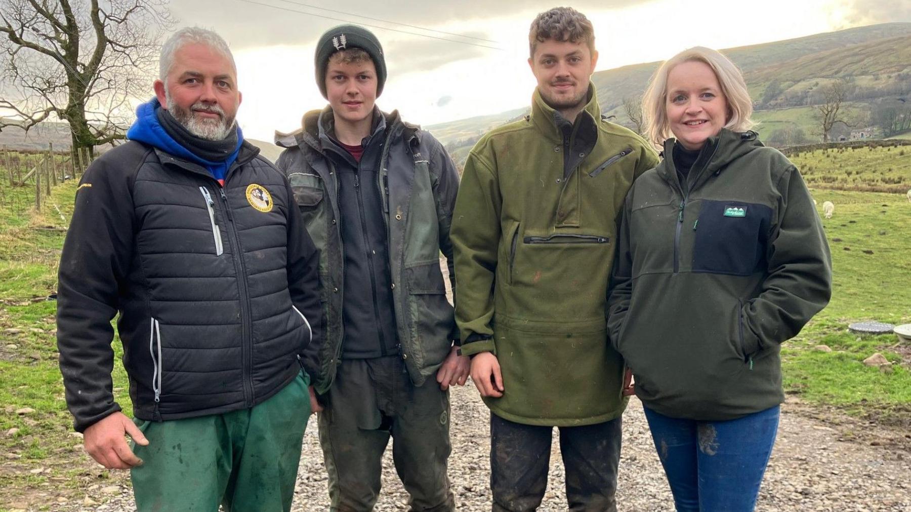 Family (left to right) Matthew Staley, Lewis Staley, Luke Staley, Liz Staley. They are standing in a field. Matthew has a grey beard and a black jacket, Lewis is wearing a black jacket and a hat, Luke is wearing a green jacket and Liz is wearing a black jumper.