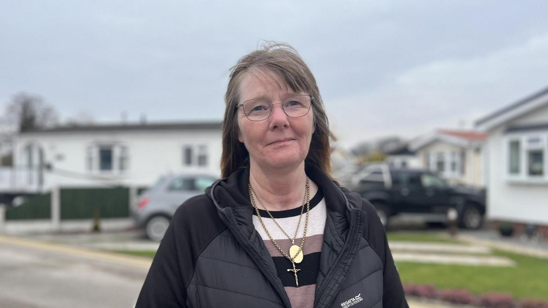 A middle-aged woman with light brown hair and metal framed glasses wearing a black puffer jacket over a black, white and brown striped jumper. She has gold chains around her neck, including a locket and a cross. The background is blurred but she is standing in front of some mobile homes with parked cars.