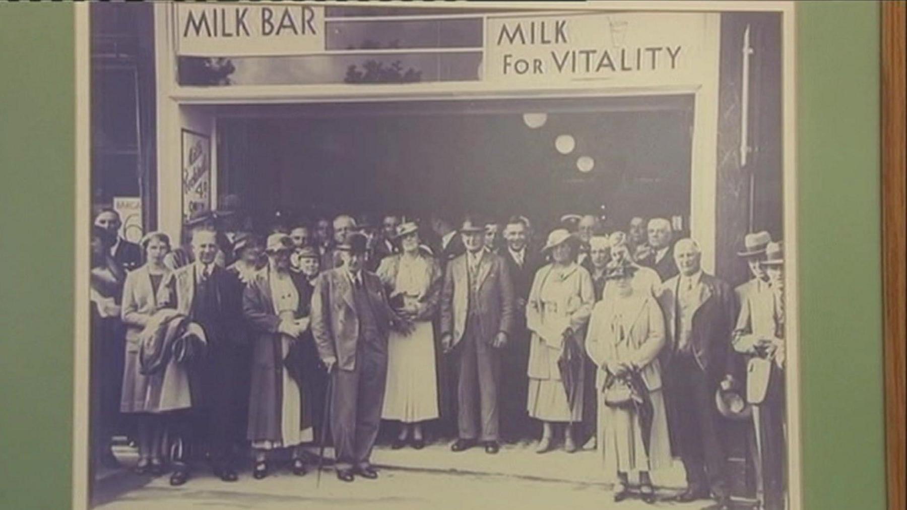 A crowd of well dressed people stand outside a milk bar 