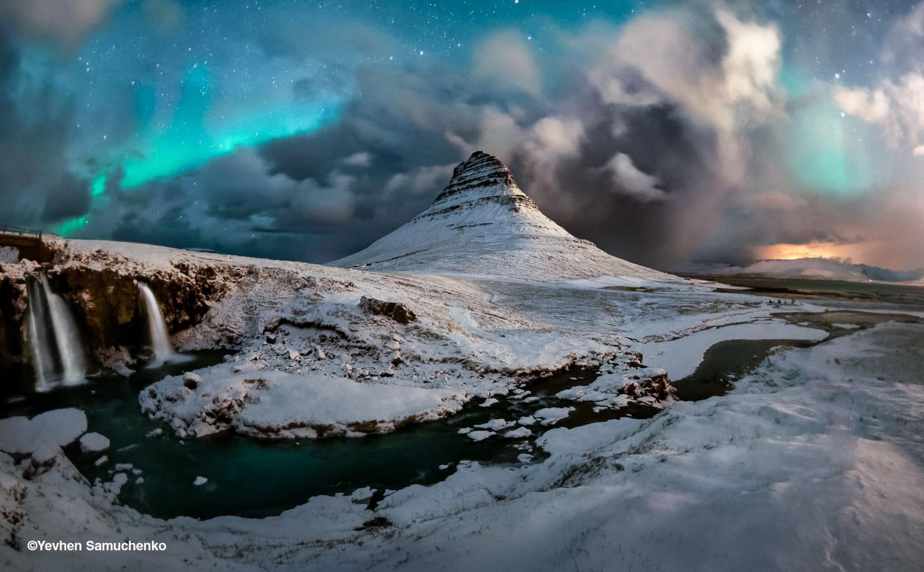 On the left of the oicture, Kirkjufellsfoss waterfall surges through snow and rock; above, the sharp mountain peak of Kirkjufell is framed by clouds. While the aurora borealis shimmers with blues and greens, with stars glinting through the gaps. 