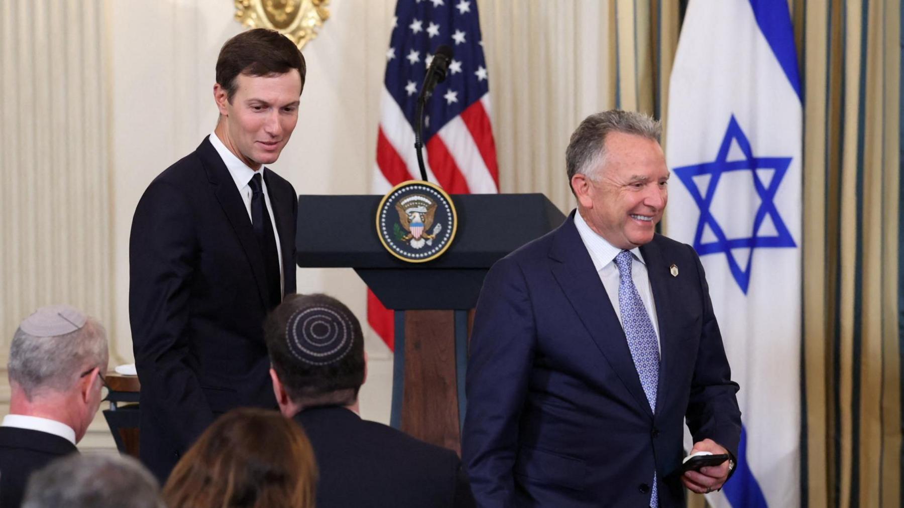 Jared Kushner, wearing a dark suit and tie, and Steve Witkoff, wearing a navy blue suit and light blue tie, walk through a press conference. A US and Israeli flag are in the background.