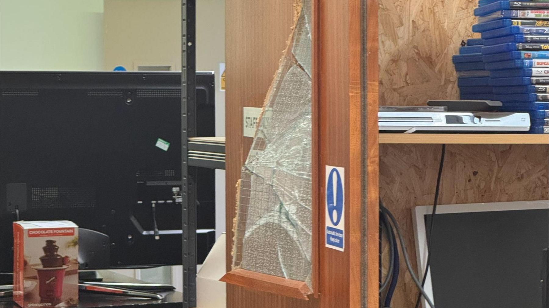 A broken window, in a charity show, showing DVDs on shelves to the right, a computer monitor to the left and right, and a hot chocolate machine in the desk.