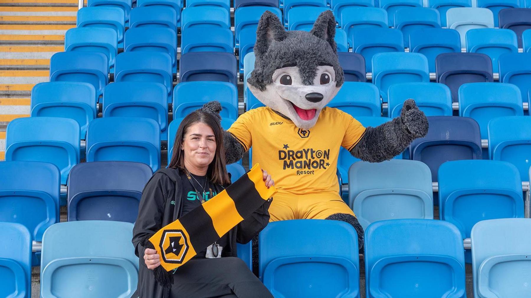 A wolf mascot in a Wolverhampton Wonderers kit is sitting behind a woman holding a black and yellow Wolverhampton Wanderers scarf. They are sitting amid rows of blue flip-up seats.