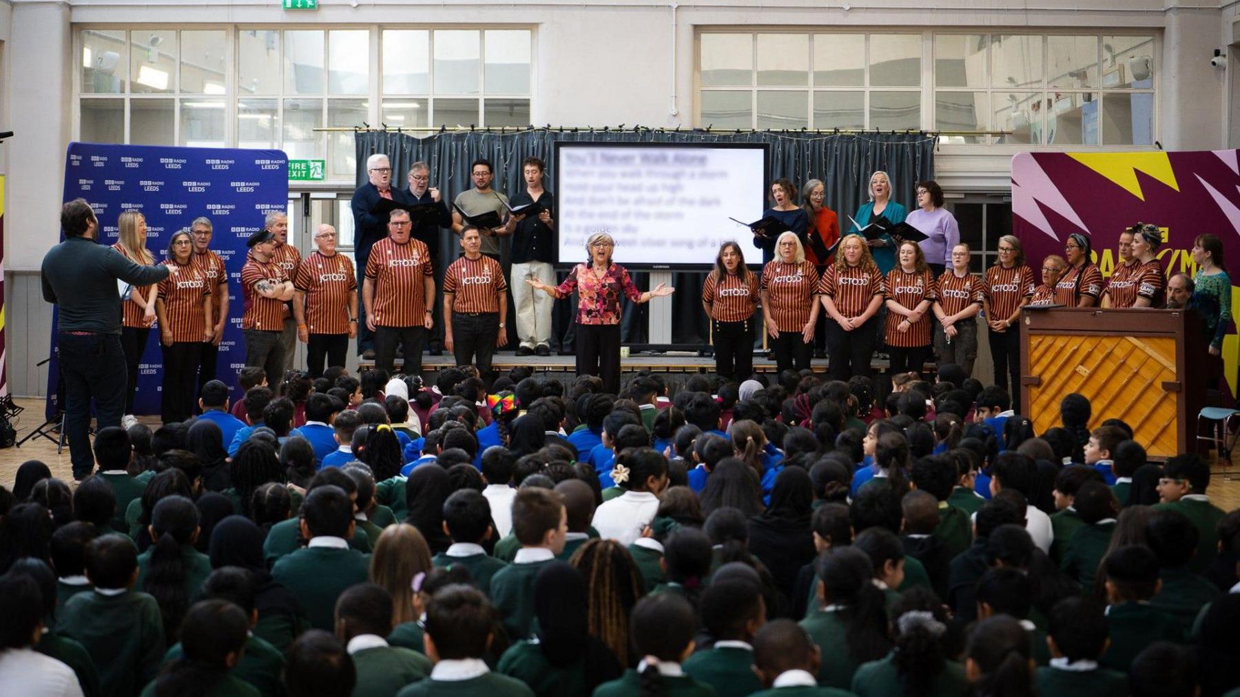 A choir on stage in front of dozens of school pupils, with the choir wearing Bradford City kits. A conductor is on the right leading the performance.