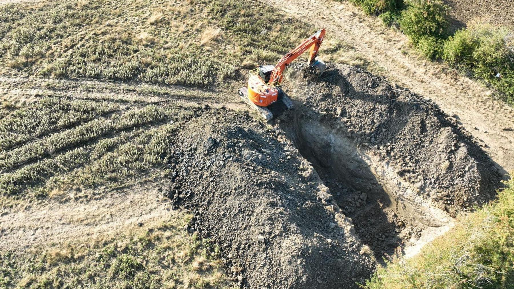 An aerial view of a digger creating a large hole in a field. There are mounds of earth either side of the hole.