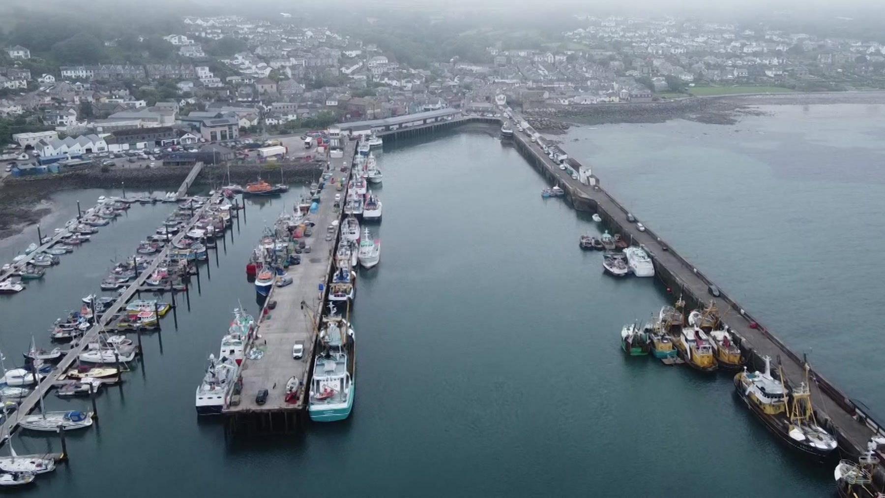 A drone shot of a harbour full of boats on a misty day. The water is a dark blue colour and there is mist and fog covering homes and the nearby town on the main land. 