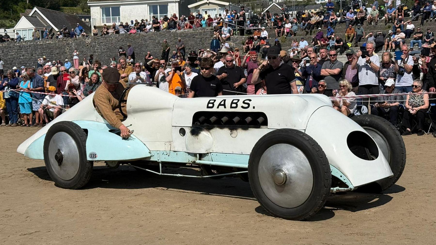 A general view of the car 'Babs' with a crowd of spectators behind it. It is white and light blue with 'Babs' written on the side in large letters. 
