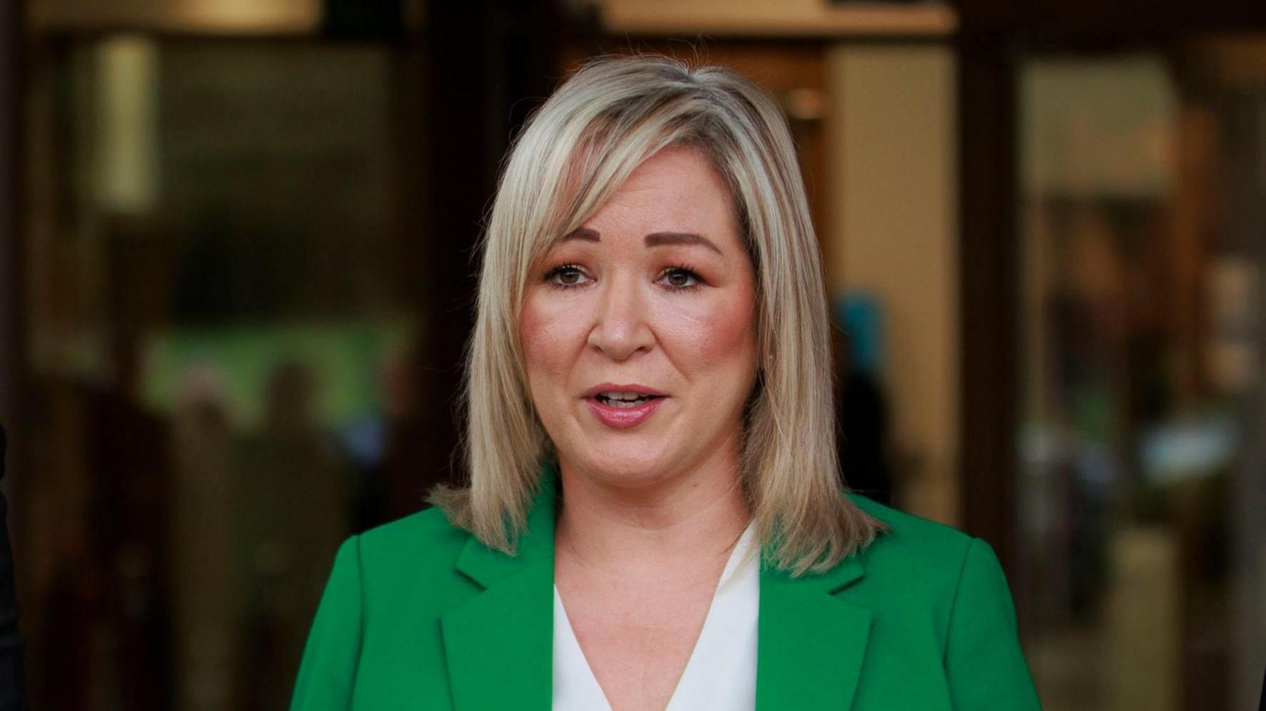 Michelle O'Neill. She has shoulder length blonde hair, wearing a green blazer and white top.