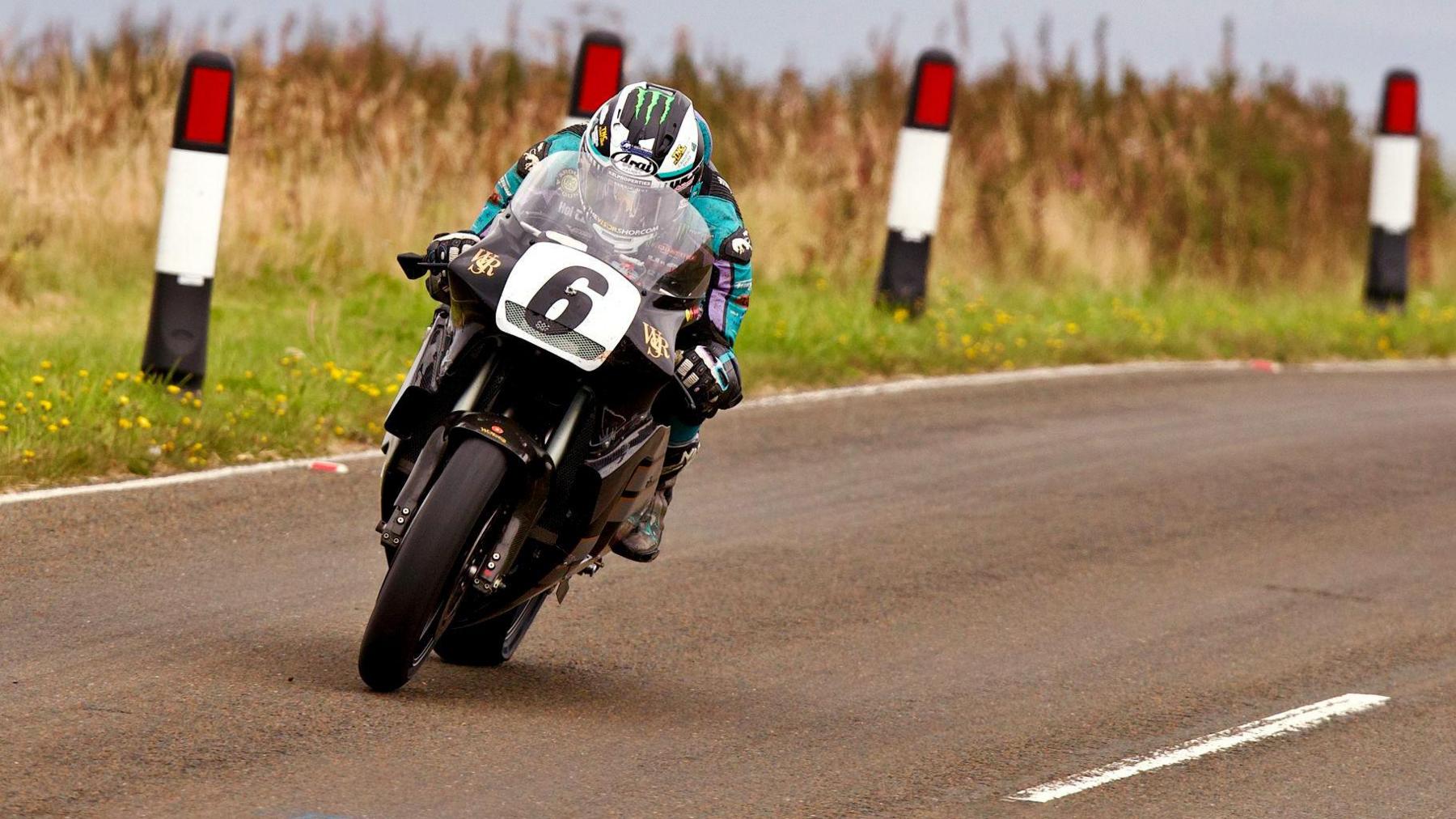 Michael Dunlop riding over the mountain on a black classic bike with the number six on the front. His leathers are coloured black, purple and turquoise.