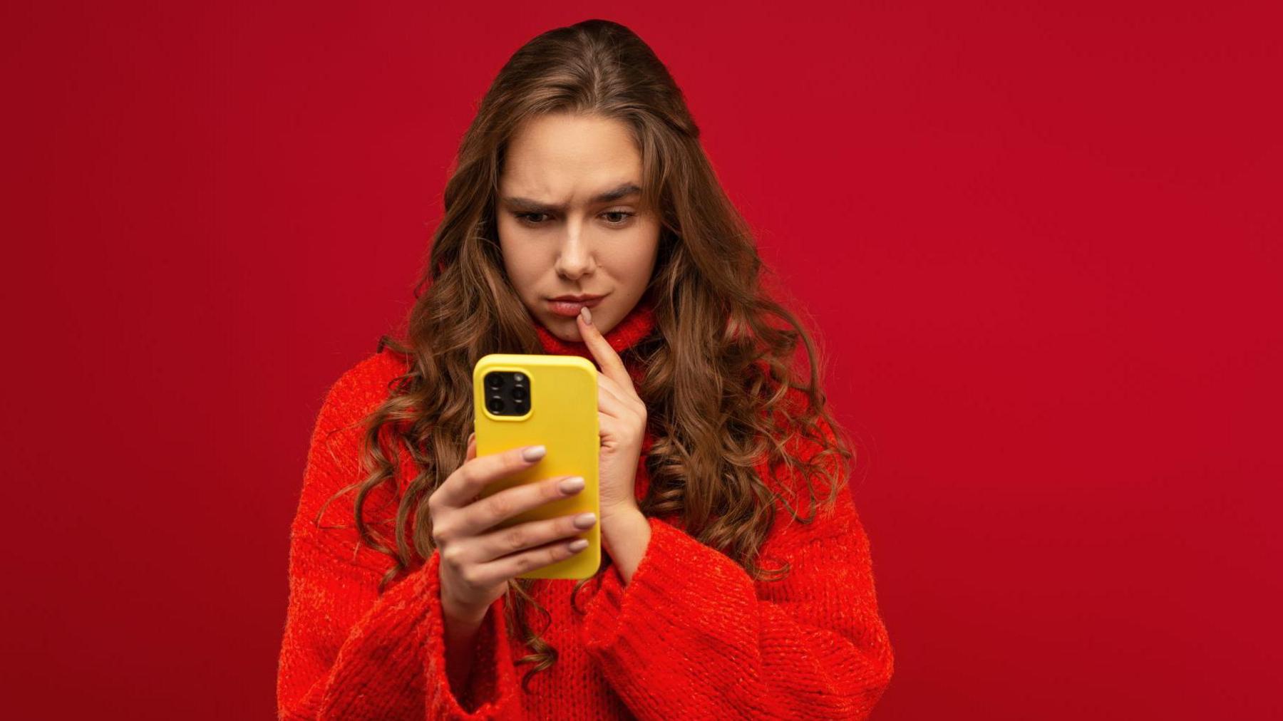 A woman in a red jumper looks quizzical as she stares at her yellow mobile phone.