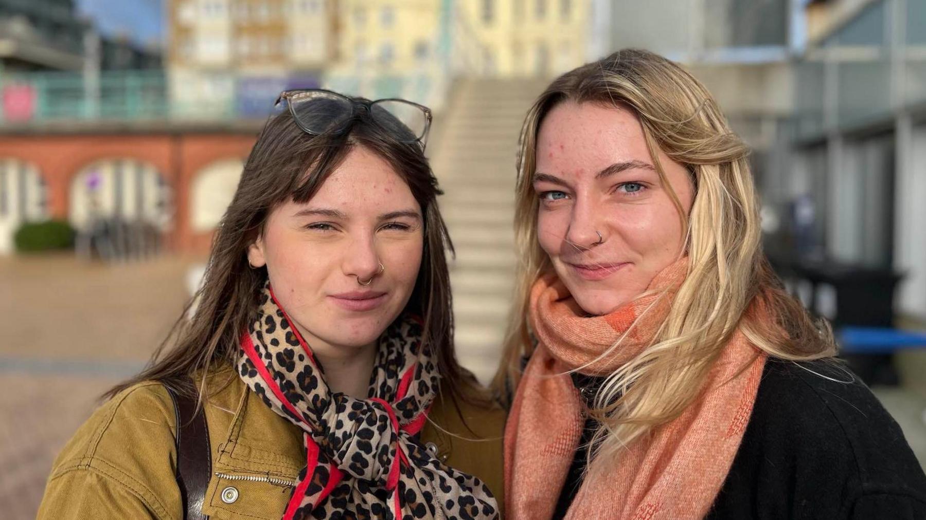 Two women - one with black hair and a leopard print scarf - and another - who is blond with a coral scarf - look at the camera with steps to the entrance of the i360 tower in the background