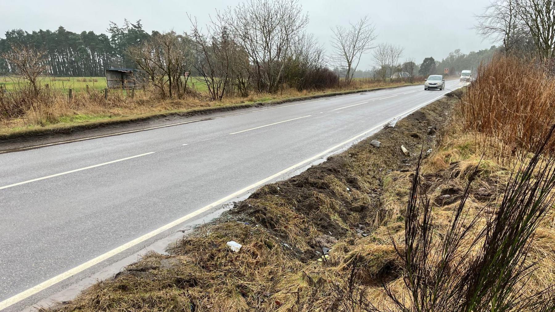 The road verge shows signs of disturbance. There is broken and flattened ground. On the road are a car and van. The sky is grey.