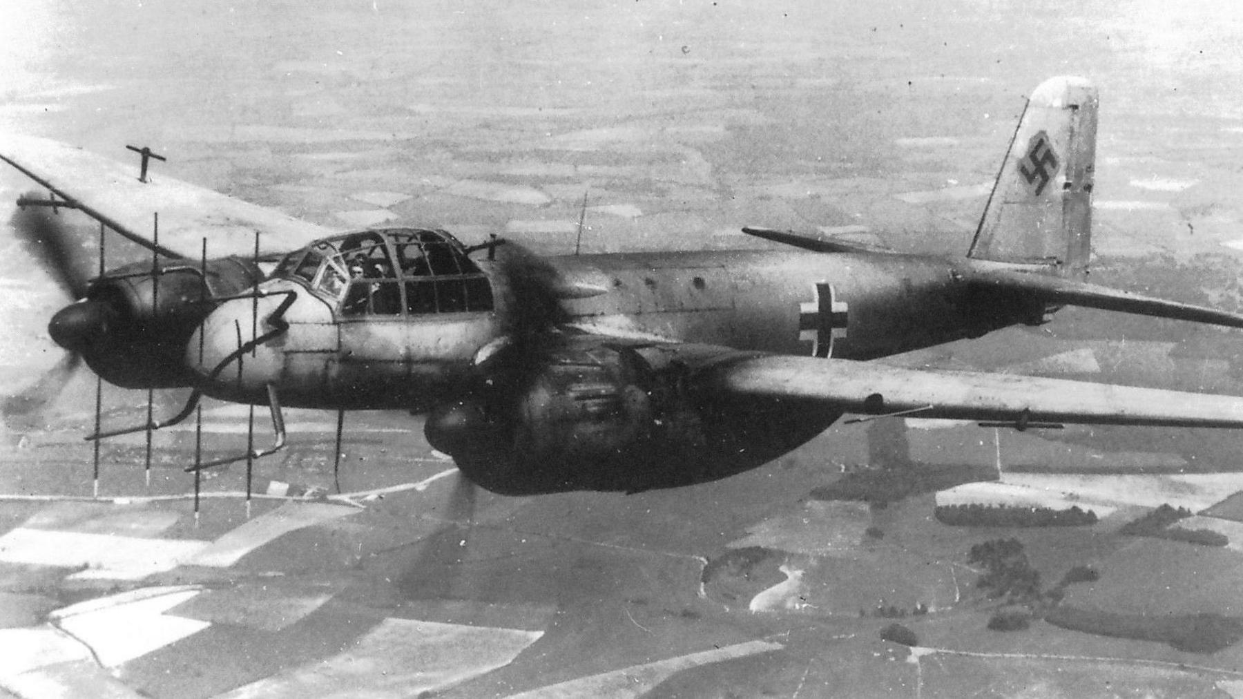 A black and white photograph of a WW2 plane flying over fields. It has two propellers and Nazi insignia 