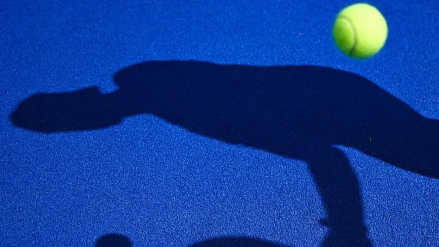 A blue padel court with a ball bouncing on the surface and the shadow of someone with a racket.