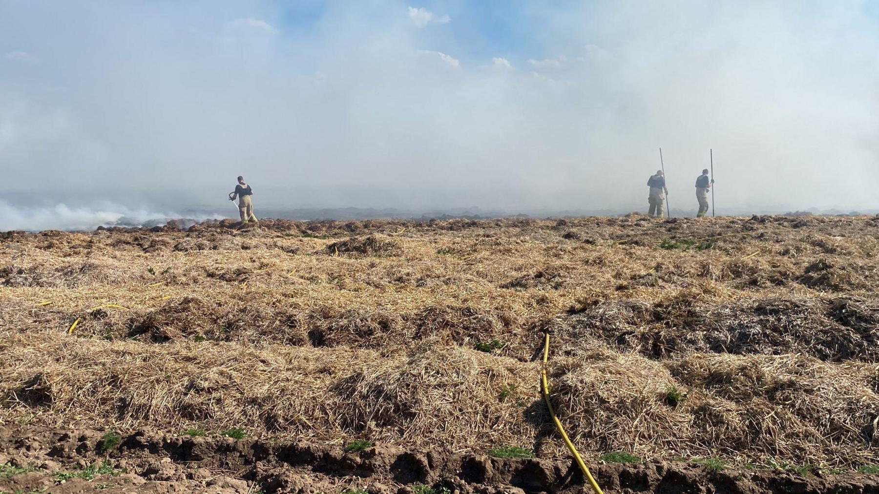 Firefighters tackle the fire in the field. Four firefighters can be seen in the picture. There is a large amount of smoke in the background.