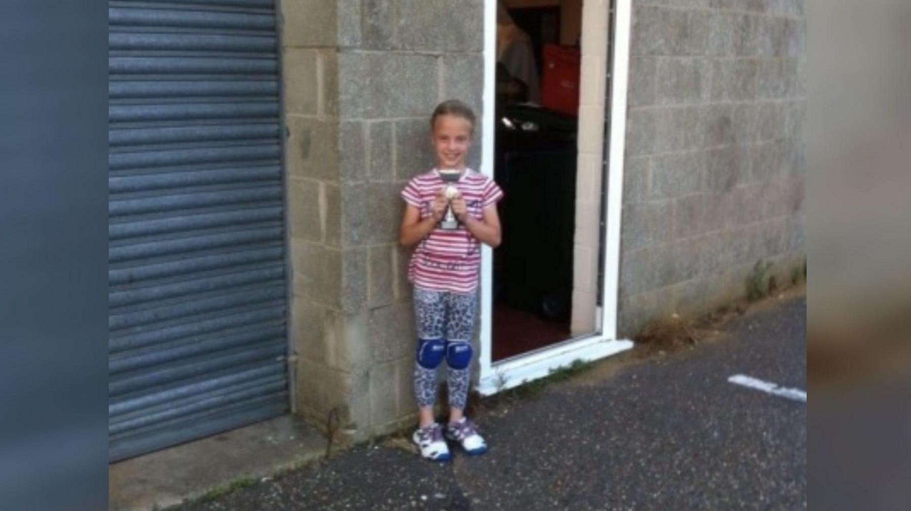 Annabelle at the age of seven holding a trophy while standing outside a building make of grey breeze blocks.