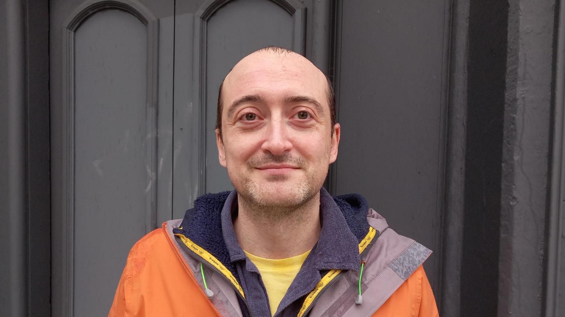 Darren John started the Save Station Street campaign after the Electric cinema shut. He has a mainly shaved head with some brown hair at his temples, stubble and wears an orange jacket with a grey lining over a blue top.