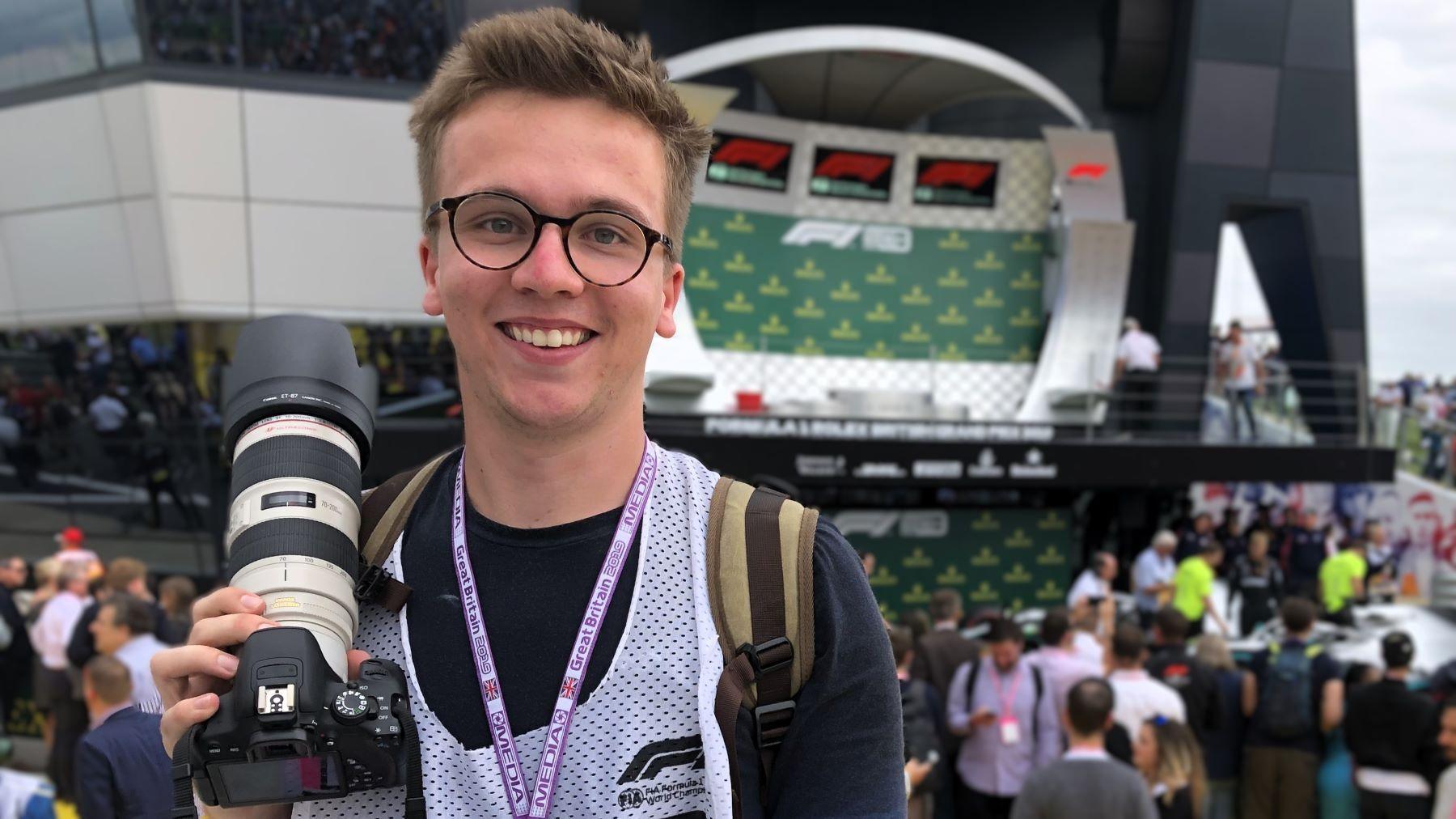 Craig Evans at a racing event holding his camera. He has spiky light brown hair and round glasses. He is stood in front of a crowd of people and he has a lanyard around his neck