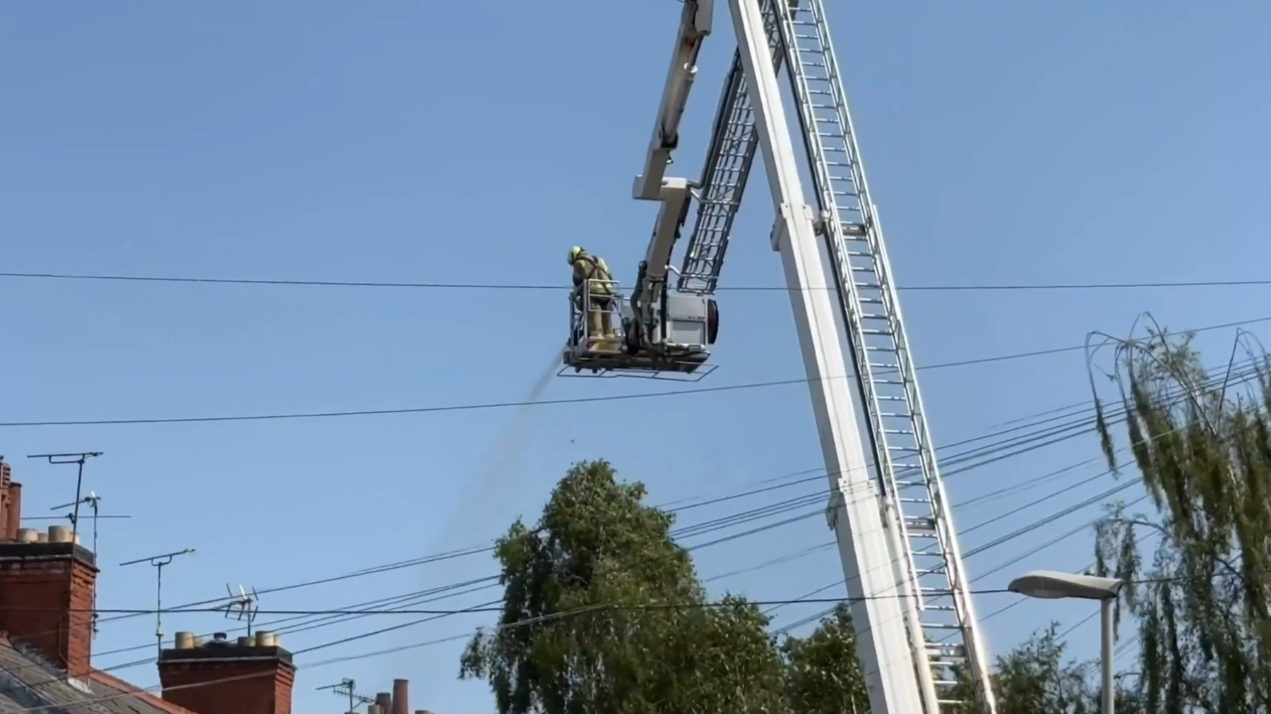 Fire crews using a hose from an elevated platform in a tree-lined street