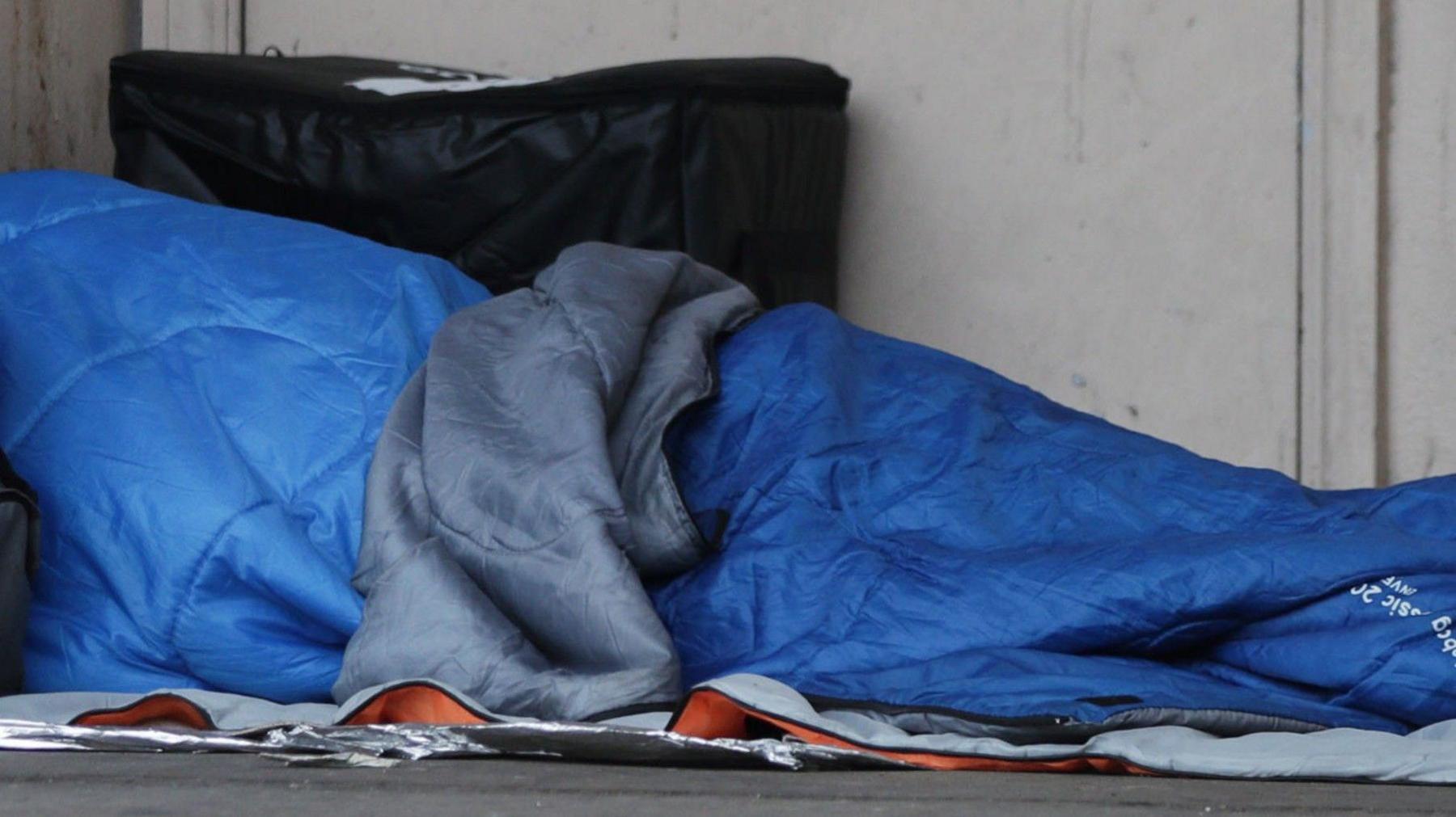 A person sleeps while covered underneath a blue and grey sleeping bag on top of newspaper.