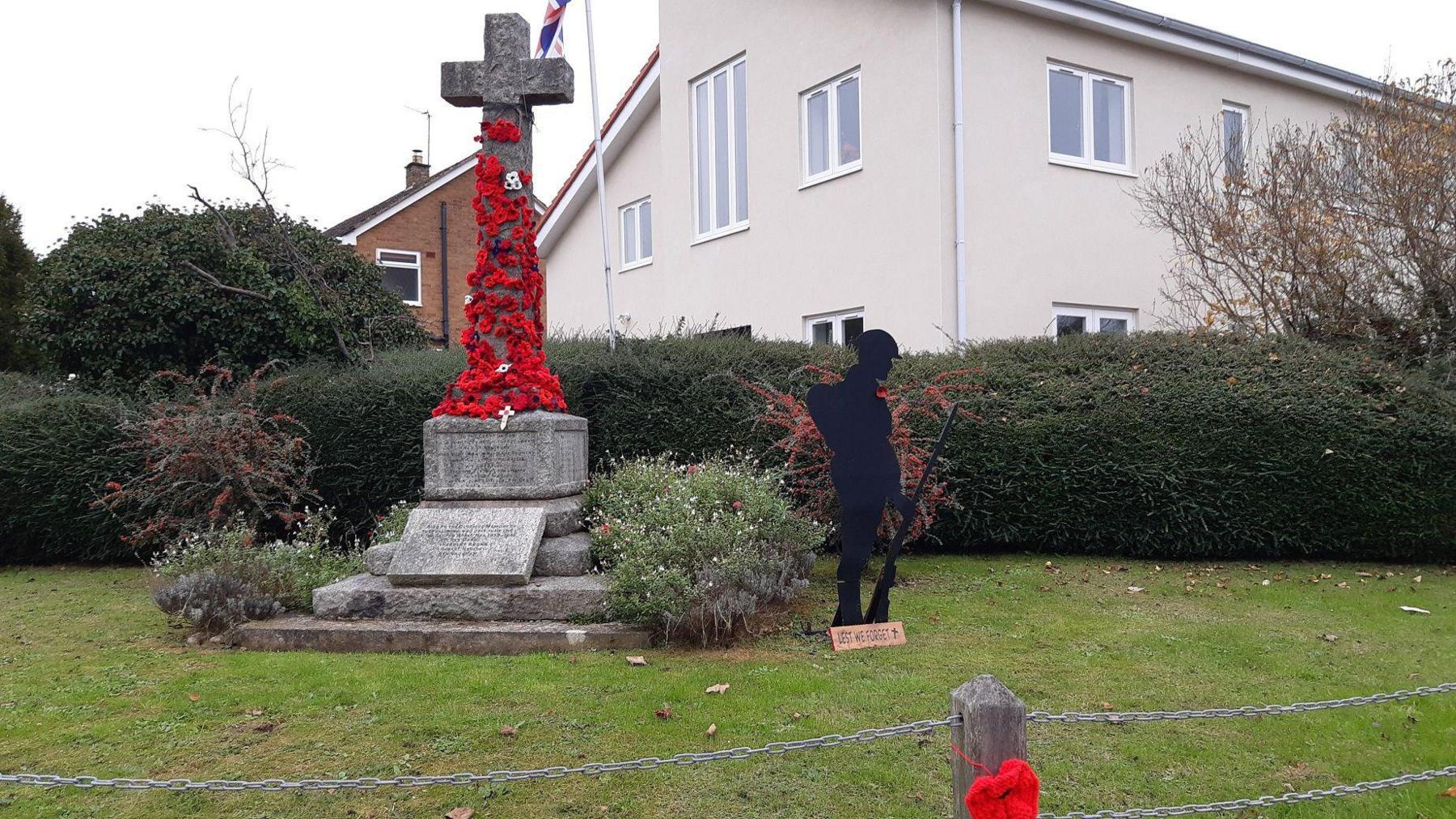 A stone water memorial stands behind a chain-link fence with a silhouette metal sculpture of a World War One soldier holding a rifle to the right of it. The memorial has been decorated with a number of red woollen poppies. Behind the memorial is a cream-coloured building with several windows with a green hedge between it and the war memorial. 