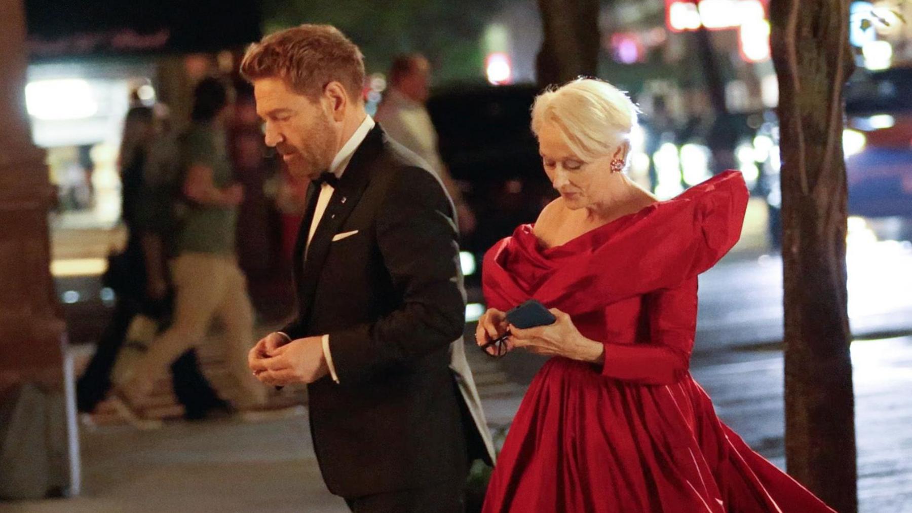 Branagh with beard in black dress suit looking pensive, with Meryl Streep in crimson red ball gown looking at her phone and holding her glasses
