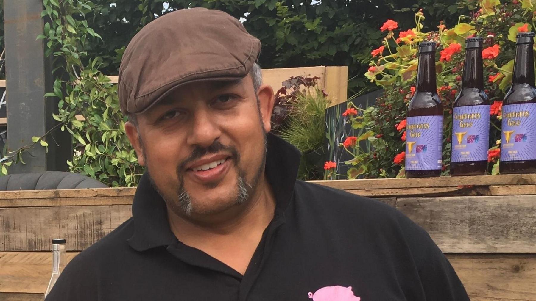 A man wearing a black polo shirt and a flat cap stands smiling in a garden.