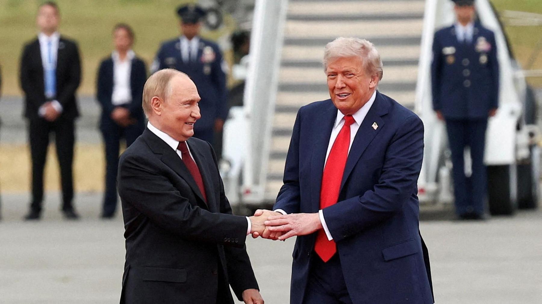 Trump and Putin stand on the tarmac in Alaska ahead of their summit, shaking hands and smiling. Behind them stand a row of uniformed men and women, and a large staircase leading up towards an aircraft. Putin is wearing a black suit, white shirt and dark red tie, and Trump is wearing a navy blue suit, white shirt and red tie. 