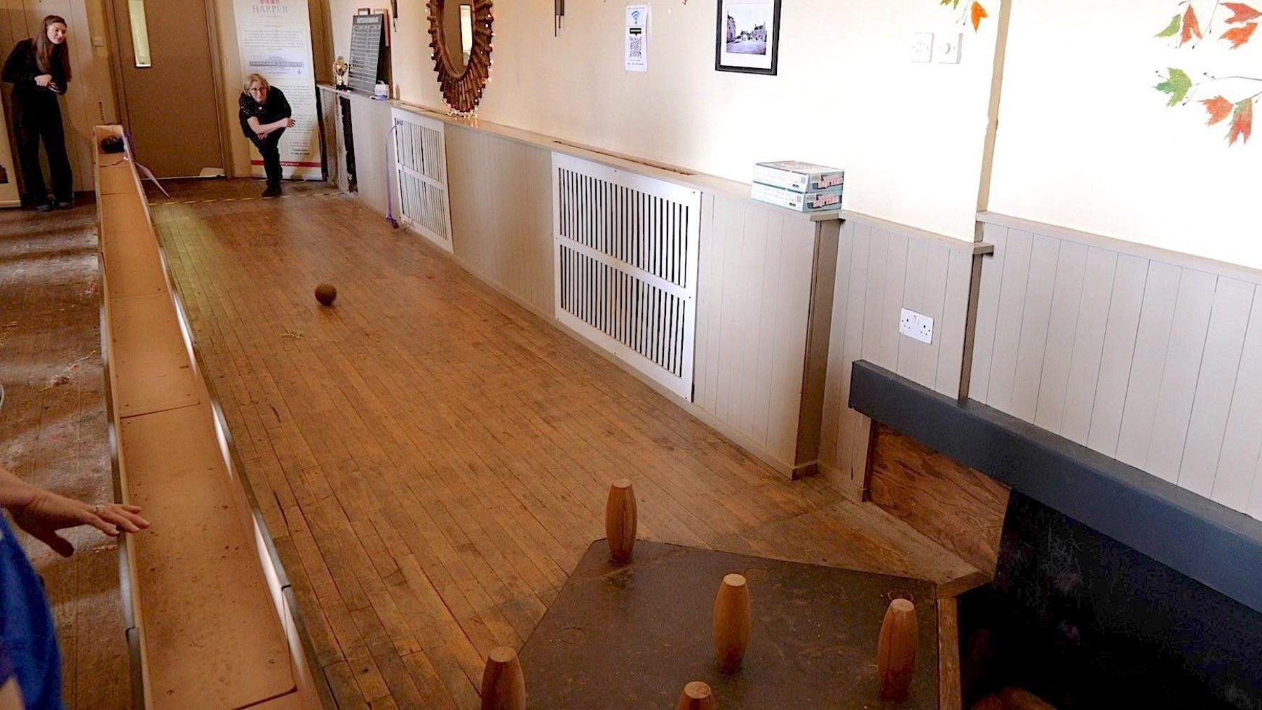 A woman wearing black bowling a skittles ball down a long wooden aisle inside a pub. 