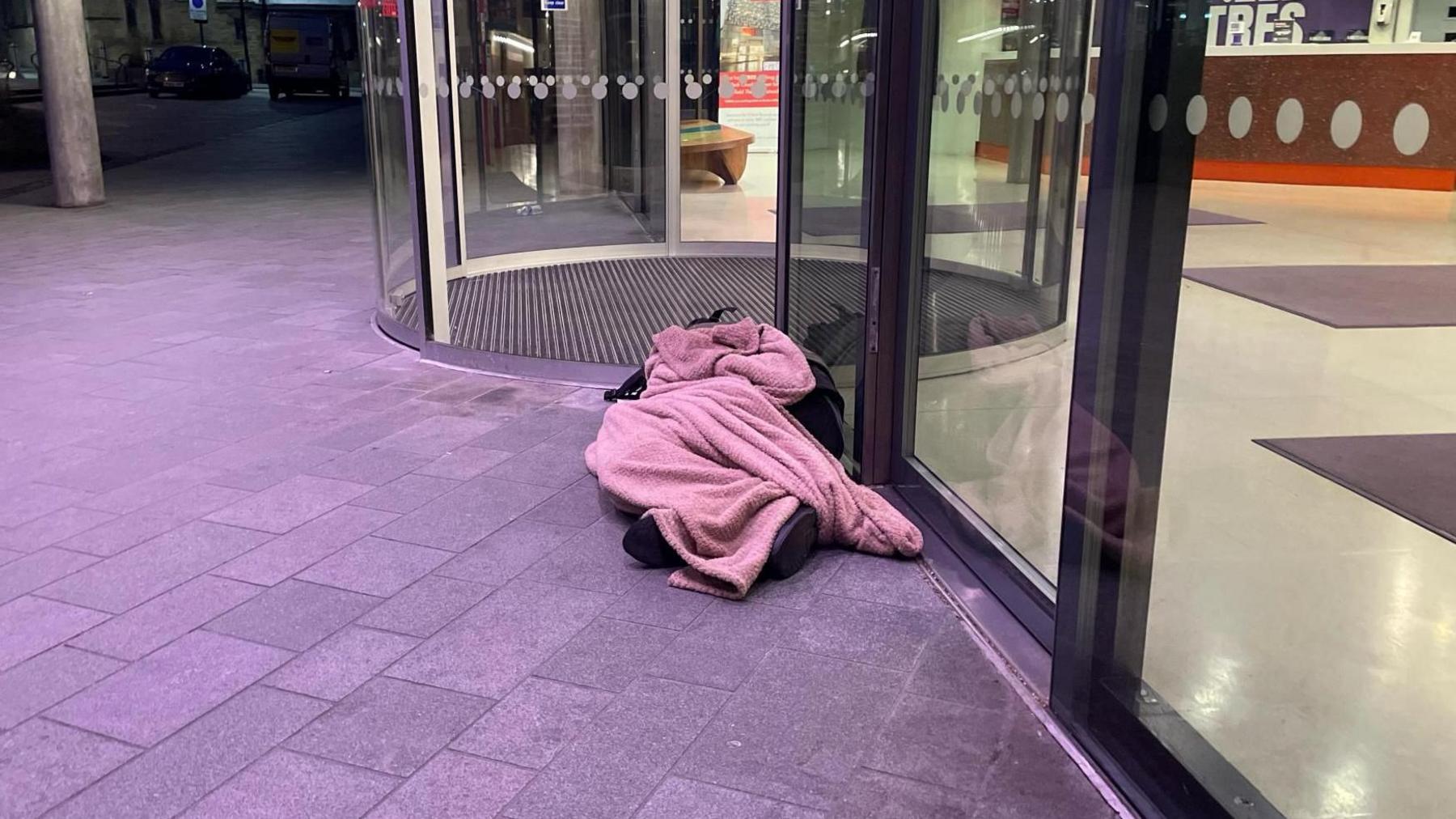 A pink blanket obscures a body underneath outside a lit building early morning