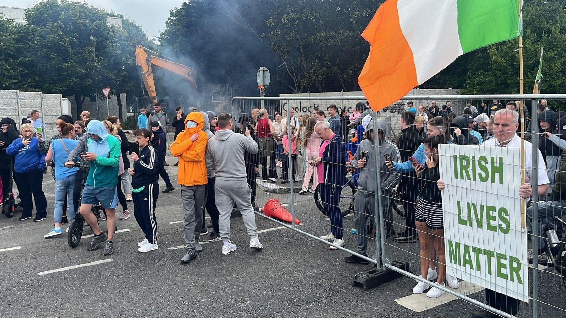 Large crowd gathers at barrier in Coolock, including one man holding a sign which reads Irish Lives Matter