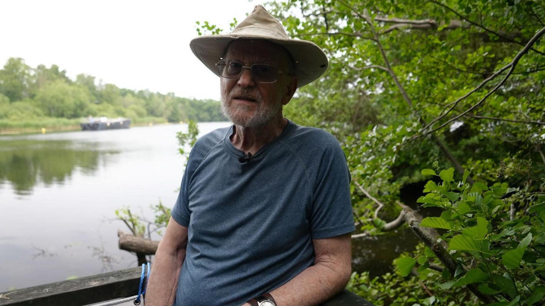 Colin Yorke is at the river's edge, surrounded by shrubbery. He wears a hat, glasses and a blue T-shirt.