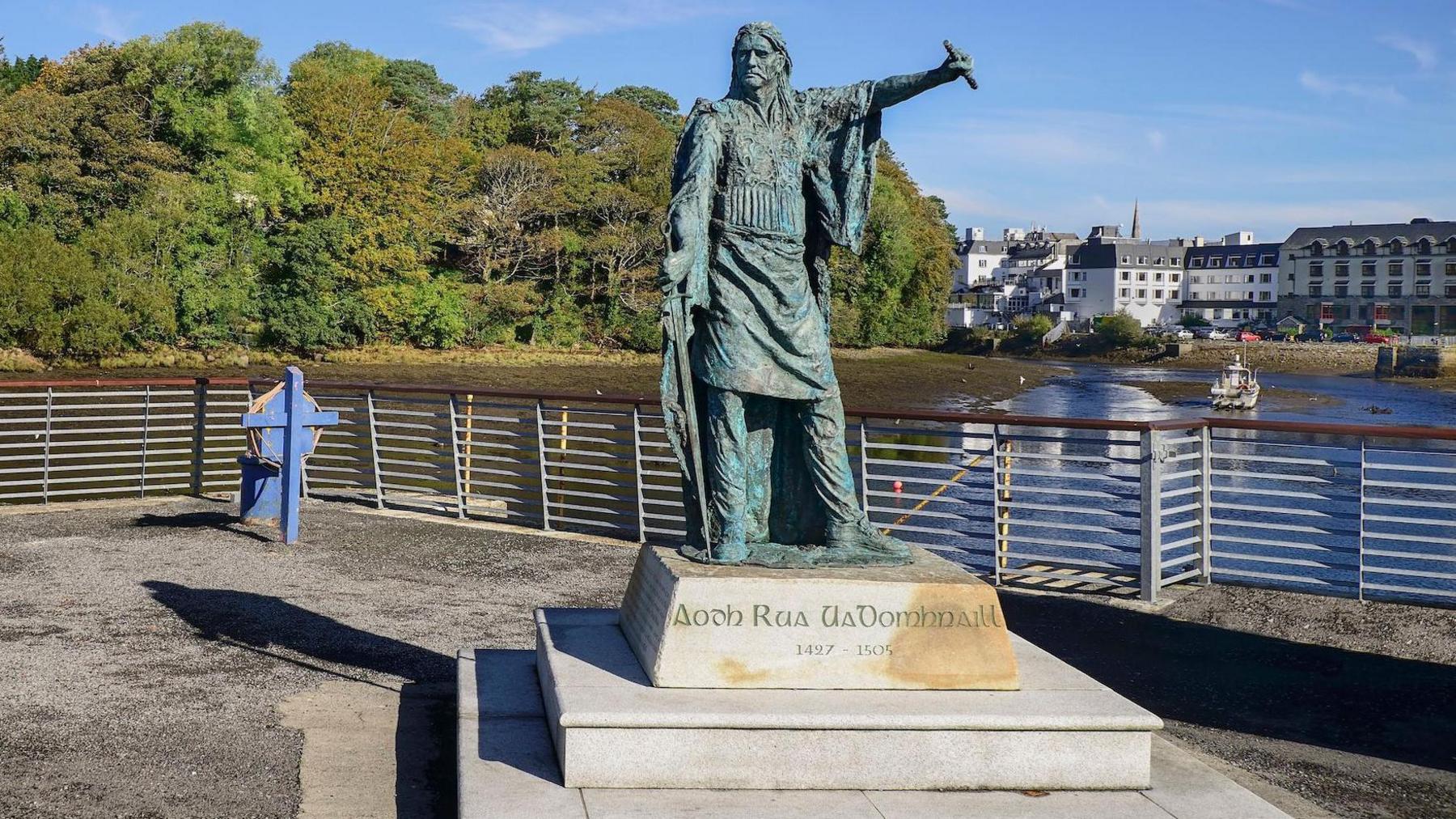 A man statue on a stone plinth sits on a concrete pathway beside a body of water. 