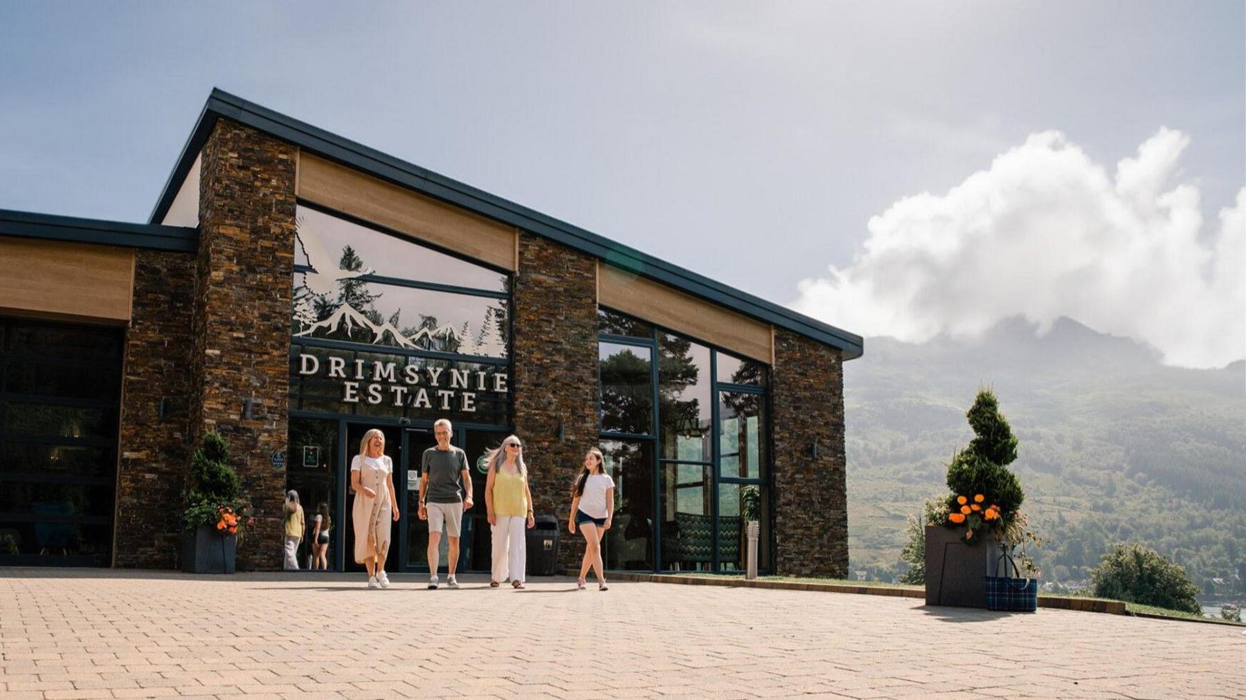 A group of people leaving a hotel with DRIMSYNIE ESTATE written in large letters above the entrance. Mountains and hills can be seen in the background.