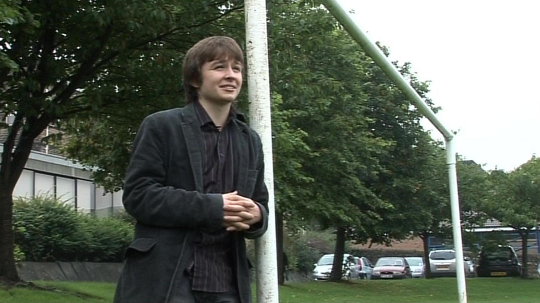 A long-haired teenager of around about 18 years of age leans against a goalpost frame