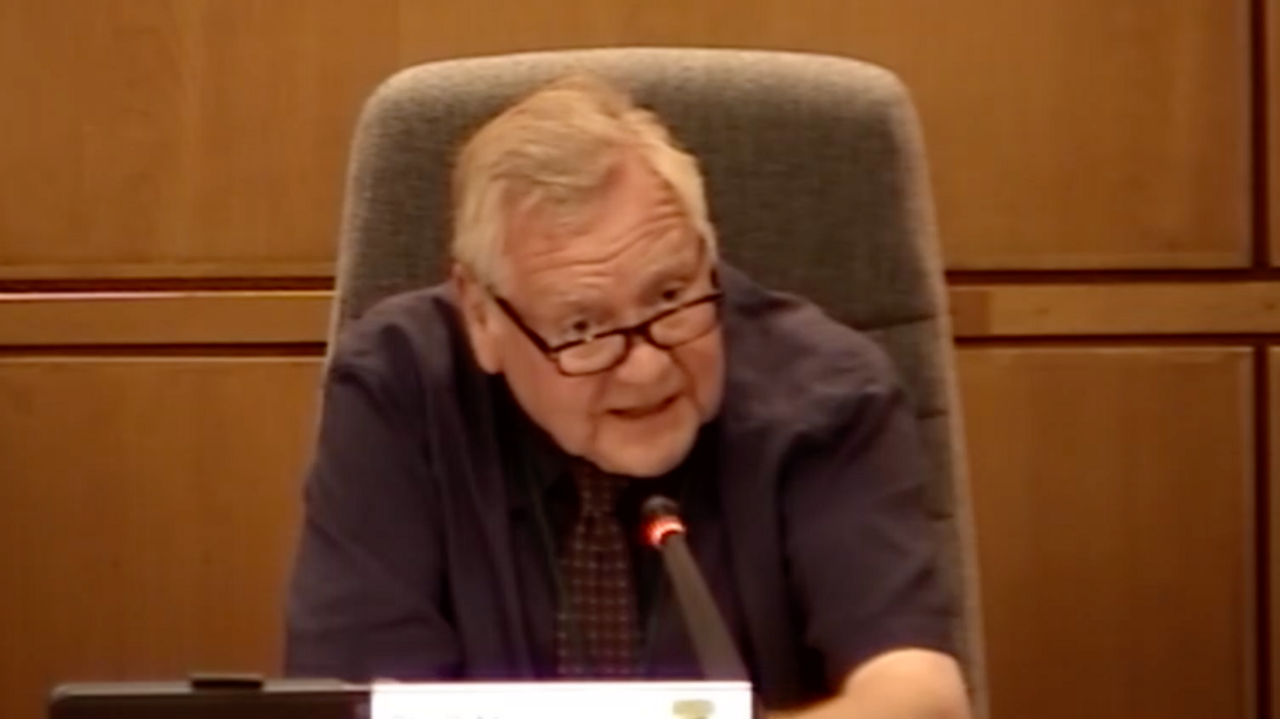 Council leader Mike Gledhill is speaking during a cabinet meeting. He is wearing a brown shirt and tie and is sitting in a brown chair with wooden panelled walls behind.