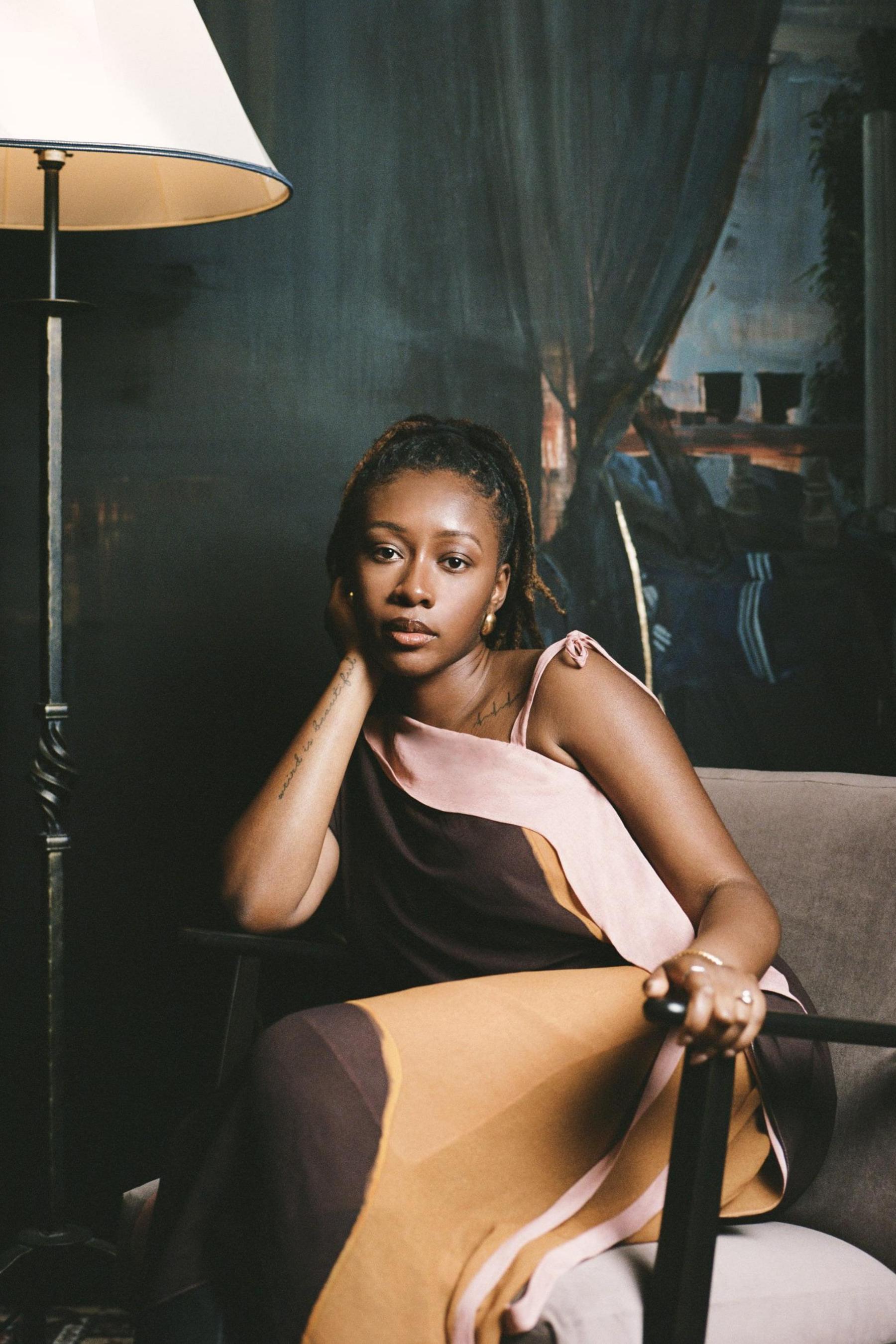 A professional portrait of the artist, a woman with afro hair pulled back into a ponytail. She is wearing a formal dress and sits on a grey chair. There is a painting behind her and a lamp to the side.